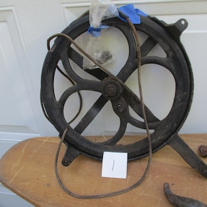 May include: An antique, cast iron wheel with a circular design and a handle. The wheel is dark gray and has a rusty appearance. A piece of wire is attached to the wheel. The wheel is sitting on a wooden surface.