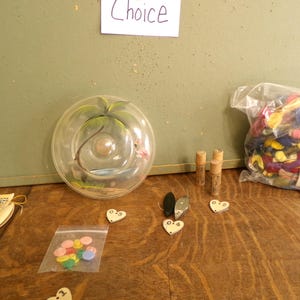 May include: A collection of craft supplies on a wooden surface. Includes a clear glass terrarium, small vials, heart-shaped tags, a bag of colorful buttons, and a bag of fabric scraps. A sign that says "Choice" is in the background.