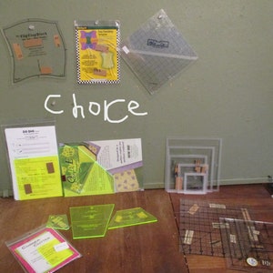 May include: Assortment of quilting tools, including clear plastic rulers, templates, and a corner cutter. The word "choice" is written in white marker on the green surface. Various sizes and shapes are visible, suitable for fabric cutting and design.