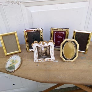 May include: A collection of decorative picture frames in various shapes and sizes. Gold, black, and white frames are displayed, including an oval frame with a floral design and a rectangular frame with a gold border. The frames are arranged on a wooden surface.
