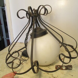 May include: Vintage chandelier with a white globe shade and ornate, dark metal scrollwork. The fixture has a chain for hanging and a decorative metal base. A small metal bracket is also visible.