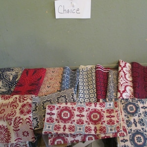 May include: A collection of folded fabric swatches featuring various patterns in red, blue, white, and black. The textiles display diverse geometric, floral, and damask-style prints. A white paper sign with the handwritten word Choice is visible.
