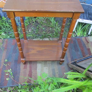 May include: A small, brown wooden side table with a rectangular top and a lower shelf. The table has four decorative, turned legs. The table is set outdoors on a multi-colored rug.