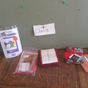 May include: A collection of craft supplies, including fabric bundles, patterns, and an applique kit. The items are arranged on a wooden surface, with a note that says "Choice" pinned to the wall in the background.