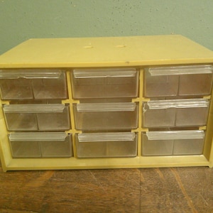 May include: A beige plastic storage cabinet featuring nine individual drawers arranged in a three-by-three grid. Each drawer is constructed from clear, transparent plastic and includes a small handle along the top edge for easy access.