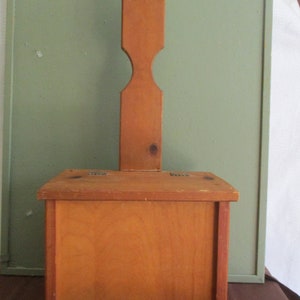 May include: A wooden butter churn with a rectangular box base and a tall, paddle-like handle. The wood has a warm, natural tone. The handle has a hole at the top. The churn is likely used for making butter.