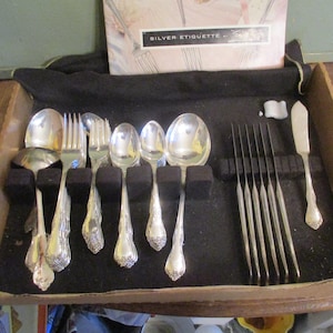 May include: A set of silver silverware, including forks, spoons, and knives, arranged in a black velvet-lined box. The set includes a serving spoon and a butter knife. The box also contains a booklet titled "Silver Etiquette."