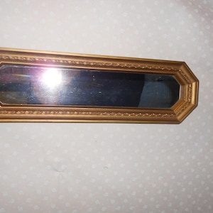 May include: A rectangular mirror with a gold-colored, octagonal frame. The frame has a decorative, textured border. The mirror is long and narrow, suitable for a hallway or bathroom. The mirror reflects the surrounding environment.