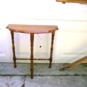 May include: A semi-circular wooden console table with a smooth, polished surface and a scalloped edge. The table features three slender, turned legs connected by a horizontal support structure, showcasing a warm, medium-brown wood grain finish.
