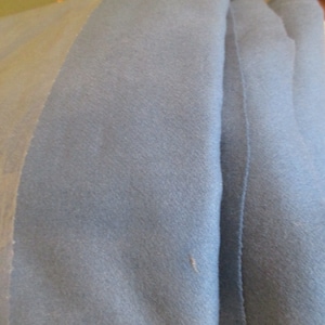 May include: Close-up of folded blue fabric. The fabric has a soft texture and appears to be a medium weight. The color is a muted blue-gray. The edge of a lighter-colored fabric is visible on the left side. A small piece of paper is in the upper right corner.