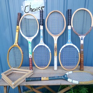 May include: A collection of six vintage tennis rackets with wooden and metal frames, featuring various string patterns and grip colors. One racket lies horizontally in the foreground, while five stand upright. A wooden press frame is also visible.