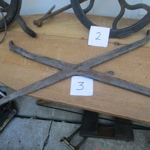 May include: An antique, rusty metal tool with an X-shaped design, possibly for a specific mechanical purpose. The tool is placed on a wooden surface, with other vintage metal objects visible in the background.