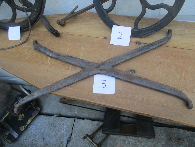 May include: An antique, rusty metal tool with an X-shaped design, possibly for a specific mechanical purpose. The tool is placed on a wooden surface, with other vintage metal objects visible in the background.