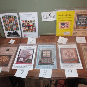 May include: Ten quilt pattern cards with various designs, including a fractured flag, a heart blossom, and a liberty homestead. The cards are on a wooden table with handwritten numbers 1 through 10 on white paper.