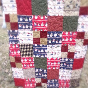 May include: A patchwork quilt with a festive winter theme. The quilt features a variety of fabrics in red, blue, green, and beige, with patterns including snowmen, plaid, and textured designs. The quilt is likely handmade and suitable for a bed or as a decorative throw.