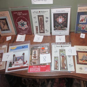 May include: A collection of 10 cross-stitch patterns featuring various themes, including a tree skirt, a snowman, a Santa, and a golfer. The patterns are displayed on a wooden table with numbers 1 through 10.