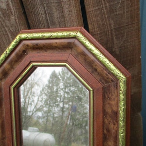 May include: Octagonal mirror with a dark brown wooden frame. The frame has a gold-colored trim and a burl wood pattern. The mirror reflects an outdoor scene. Decorative wall mirror.