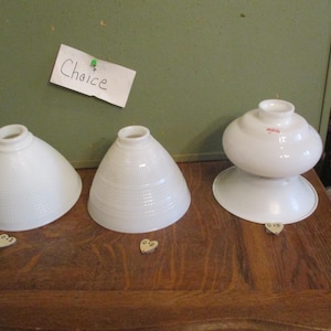 May include: Three white glass lamp shades of different shapes are displayed on a wooden surface. The shades include a cone shape, a rounded cone, and a double-layered design. A small paper with the word "Choice" is pinned to the wall.