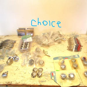 May include: A collection of various cabinet hardware, including knobs, pulls, and handles, in silver and gold tones. Some items are packaged, while others are loose. The word "Choice" is written in blue on the wall.