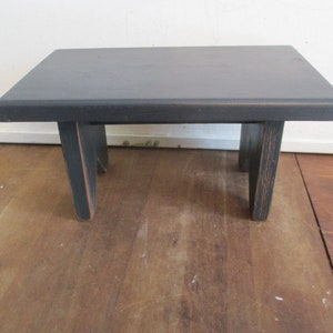 May include: A small, black wooden step stool with a rectangular top and four legs. The stool is painted black with visible wood grain and measures approximately 12 inches long, 10 inches wide, and 8 inches tall.