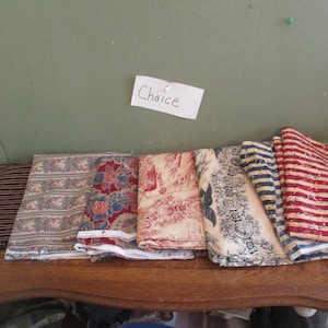 May include: A selection of fabric swatches with various patterns, including stripes, floral designs, and toile. The fabrics are displayed on a wooden surface, with a handwritten note that says "Choice" pinned above them.