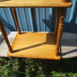 May include: A small, vintage wooden side table with a light brown finish. The table has a square top and a lower shelf, supported by four turned legs. The table is likely used for decorative purposes.