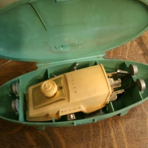 May include: Vintage Singer sewing machine in a green carrying case. The machine is beige with a gold-colored grill and a white knob. The case is open, revealing the machine and its accessories. The case has a rounded, oval shape.