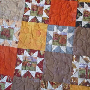 May include: Close-up of a patchwork quilt featuring squares in shades of orange, yellow, brown, and beige. Some squares have star patterns, while others have quilted circle designs. The quilt has a rustic, autumnal theme.