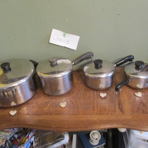 May include: Four stainless steel pots with lids and black handles are arranged on a wooden surface. The pots vary in size, with the largest on the left. Small heart-shaped decorations are placed below the pots.
