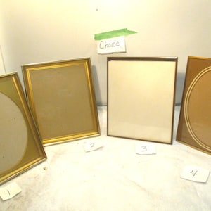 May include: Four picture frames in various shapes and sizes. Two have oval openings, and two are rectangular. The frames have gold and bronze tones. The word "Choice" is written on a small piece of paper taped to the wall.