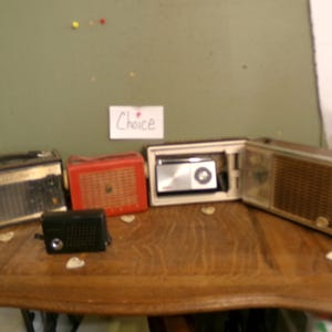 May include: A collection of vintage radios in various colors and designs, including a black, a red, and a silver radio. The radios are displayed on a wooden table. A small sign reads "Choice".