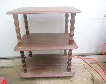 May include: A three-tiered wooden side table with a dark brown finish. The table has turned legs with a decorative design and three shelves.