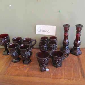 May include: A collection of ruby red glassware, including goblets, mugs, small cups, and two candlesticks. The glassware features a grape design. A small sign reads "Choice".