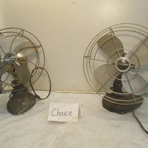 May include: Two vintage electric fans with metal blades and protective cages. One fan has a gold-toned base and the other has a black base. The fans are displayed on a white surface. The word "Choice" is written on a piece of paper.