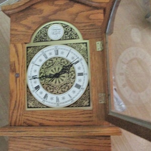 May include: A wooden wall clock with a light brown finish. The clock face is white with black Roman numerals and ornate gold detailing. The clock has a glass door and a decorative top with the text "Tempo Fugit".