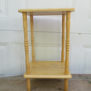 May include: A small, light-colored wooden side table with two rectangular shelves. The table has four turned legs and measures approximately 24 inches tall. The wood has a natural finish, showcasing the grain.