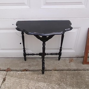 May include: A black semi-circular console table featuring a scalloped edge tabletop, three slender turned legs, and a central decorative support bracket connecting the legs beneath the tabletop surface, finished in a solid matte black paint.