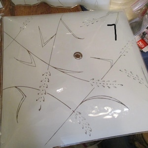 May include: Square, off-white glass ceiling light fixture with a wheat stalk design in brown. The fixture has a central circular opening and is surrounded by a thin, gold-colored trim. The number "7" is written in black on the glass.
