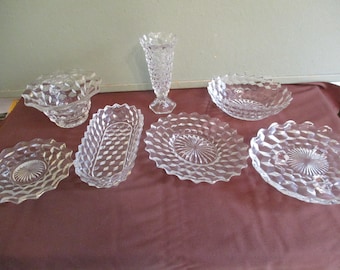 May include: A collection of clear, diamond-patterned glass serving dishes and a vase. The set includes a small bowl, a rectangular dish, several plates with scalloped edges, and a tall vase. The pieces are arranged on a maroon surface.