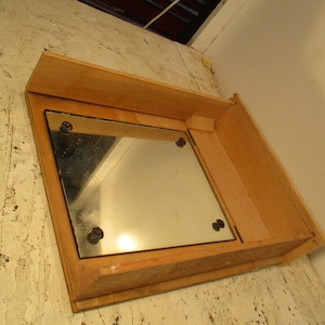 May include: A wooden framed mirror with a square shape. The mirror is set into a wooden frame with a small shelf. The mirror is held in place by four decorative, dark-colored fasteners.