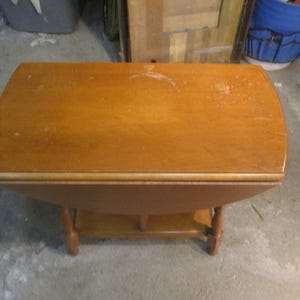 May include: A wooden drop leaf table with a light brown finish. The table has a rounded top and a lower shelf. The table is in good condition with some minor scratches and wear.