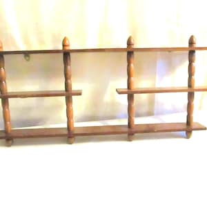 May include: A vintage wooden wall shelf with three tiers. The shelf is made of dark brown wood and features decorative turned supports. The top of the shelf has a decorative finial on each end. The shelf is designed to be mounted on a wall.