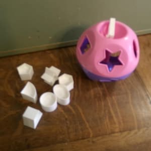 May include: A pink and purple shape sorter toy with a white handle and various shaped holes. White geometric shapes are scattered nearby. The toy is designed for early childhood development and learning.