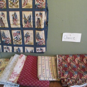 May include: Assorted patriotic fabric pieces. One piece features rectangular panels with illustrations of the American flag and figures. Other fabrics include red, white, and blue patterns, including stars, stripes, and eagles. The word "Choice" is written on a small piece of paper.