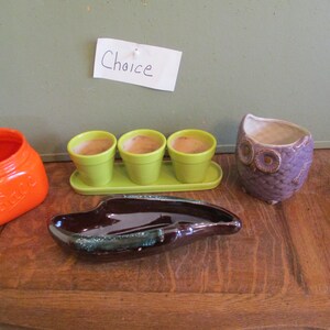 May include: An assortment of ceramic planters and containers. Includes an orange jar, three small green pots on a tray, a brown ashtray, and a purple owl-shaped planter. The word "Choice" is written on a small white paper.