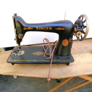 May include: An antique Singer sewing machine, black with gold accents, sits on a wooden surface. The machine features the Singer logo and decorative patterns. A brown cord is draped across the machine.