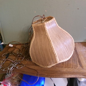 May include: A vintage lampshade with a textured, light brown surface and a cream-colored trim. The lampshade is pear-shaped and has a metal frame. A chain and electrical cord are attached.
