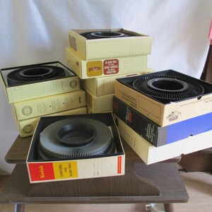May include: A stack of vintage Kodak Carousel slide trays in their original boxes. The boxes are yellow and brown with black text. The trays are black and gray with a circular opening in the center.