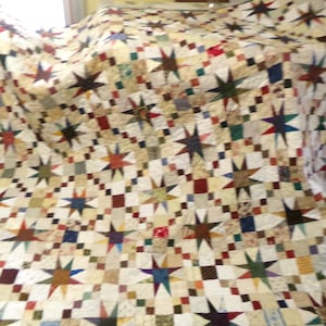 May include: A large, handmade quilt with a star pattern. The quilt features a cream-colored background with a repeating pattern of colorful stars in shades of red, blue, green, and brown. The quilt is likely made of cotton.
