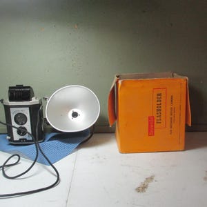 May include: Vintage black and silver box camera with a flash attachment and an orange box labeled "Flashholder". The camera has a lens, dials, and a cord. The flash has a large, silver reflector.